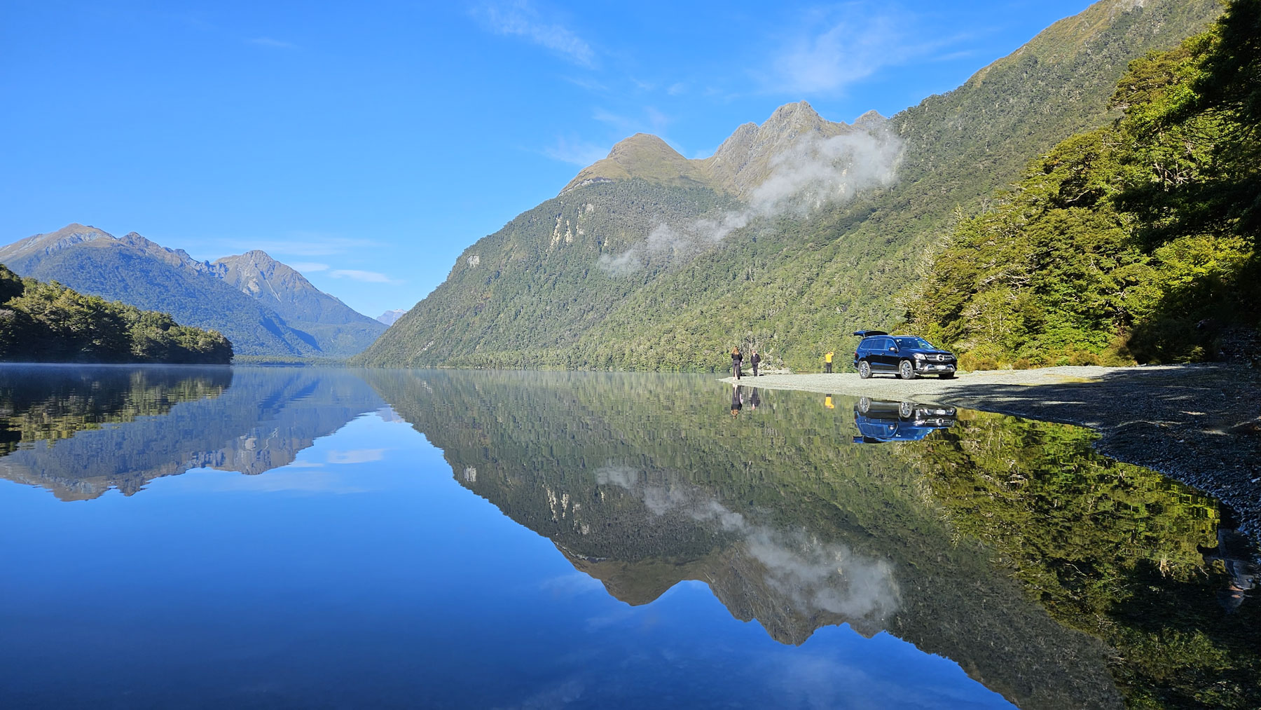A luxury car and 3 people on the shoreline of Lake Gunn with full mountain reflections