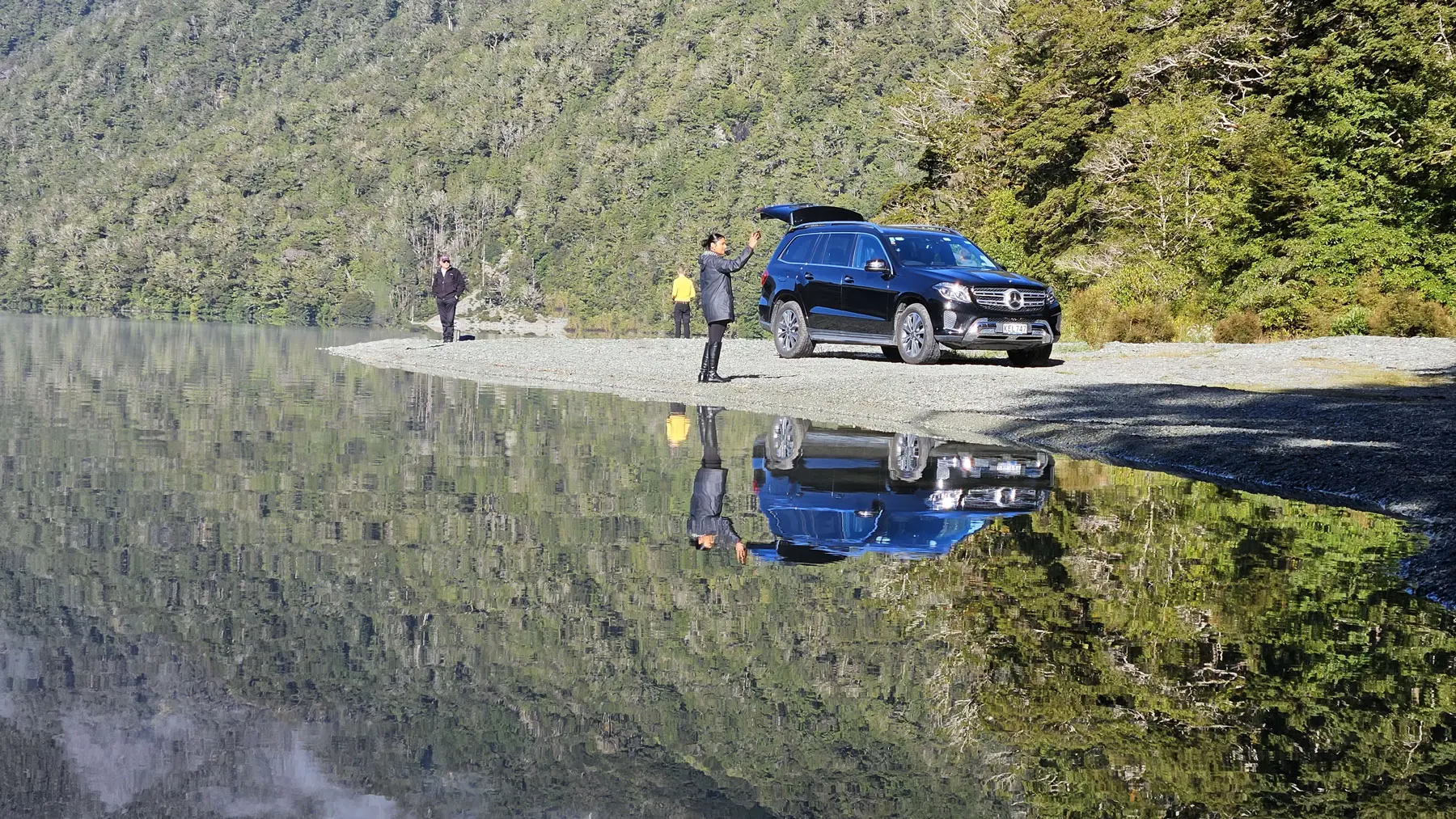 Visitors on a Lake Gunn beach showing impressive reflections with their luxury vehicle during a Milford Sound Private Tour.